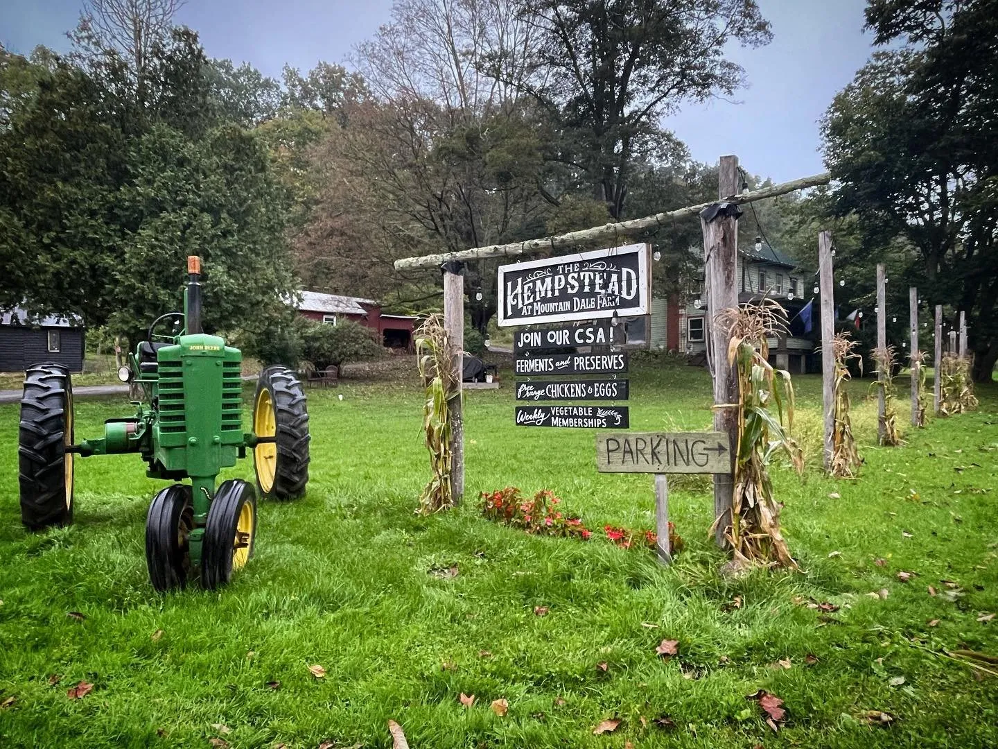 A green vintage tractor parked on grass beside a hand-painted Hempstead farm sign advertising the CSA, with autumn trees behind.