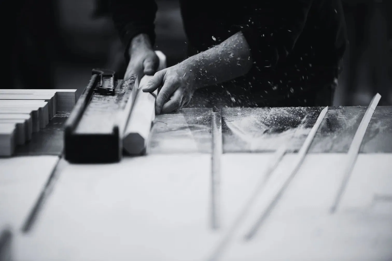 Black and white photograph of hands guiding a piece of wood through a table saw, sawdust in the air.